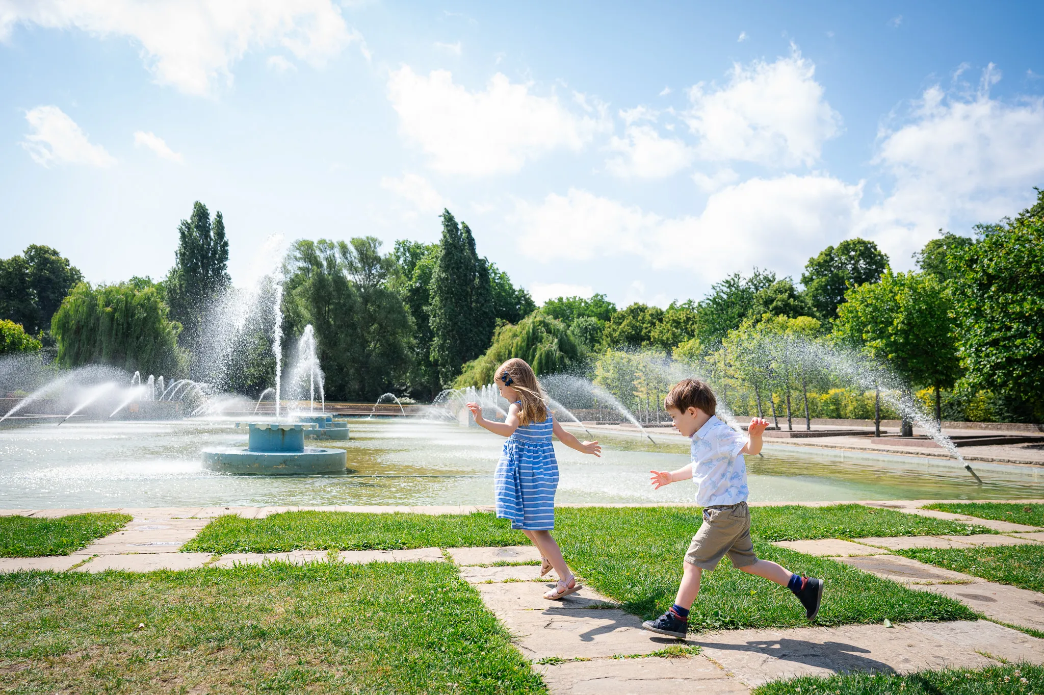 Brother and sister running and playing by the fountains in Battersea Park on a sunny day